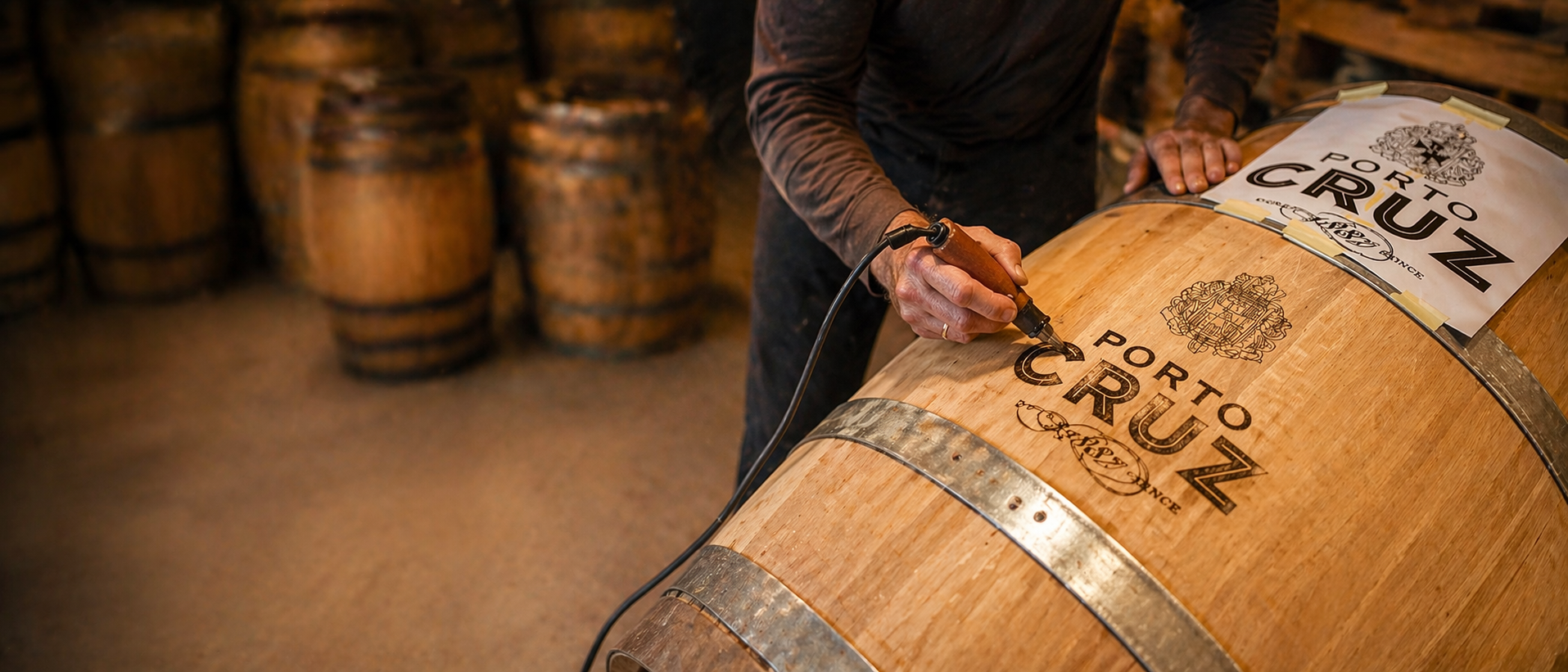 Handwerker brennt mit einem Brennwerkzeug das Logo „Porto Cruz“ präzise in ein Holzfass ein, während im Hintergrund unscharf weitere Fässer in einer warm beleuchteten Werkstatt zu sehen sind.