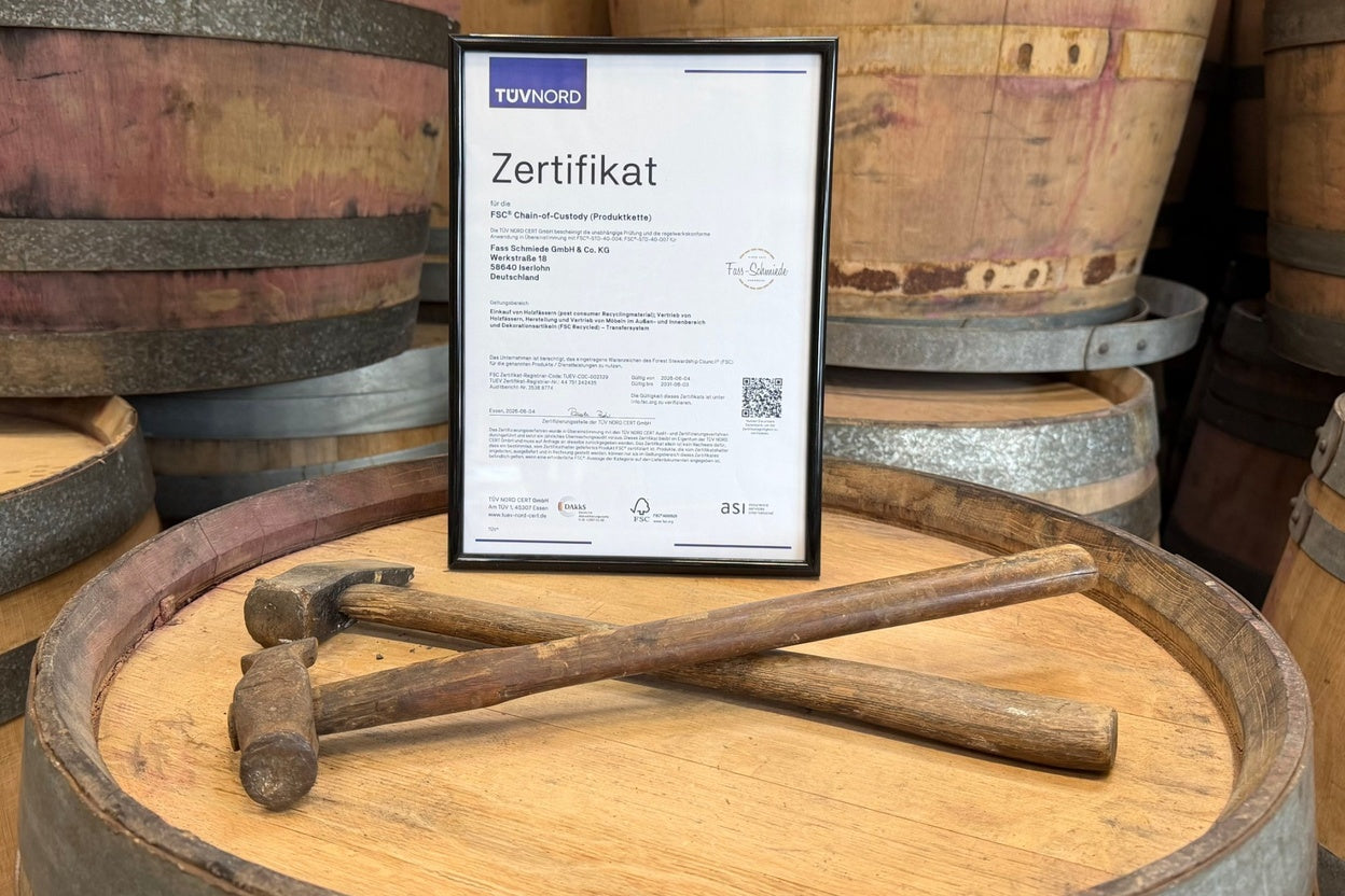 Gerahmtes FSC®-Zertifikat auf einem Weinfass, davor zwei historische Fassmacherhämmer, im Hintergrund gebrauchte Weinfässer.