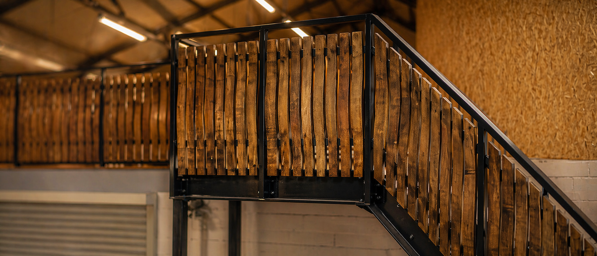 Modernes, warm ausgeleuchtetes Industrie-Interieur mit Fokus auf ein hochwertiges Treppengeländer aus Holz und schwarzem Metall; der Hintergrund ist weichgezeichnet und betont die handwerkliche Struktur der vertikalen Holzstreben.