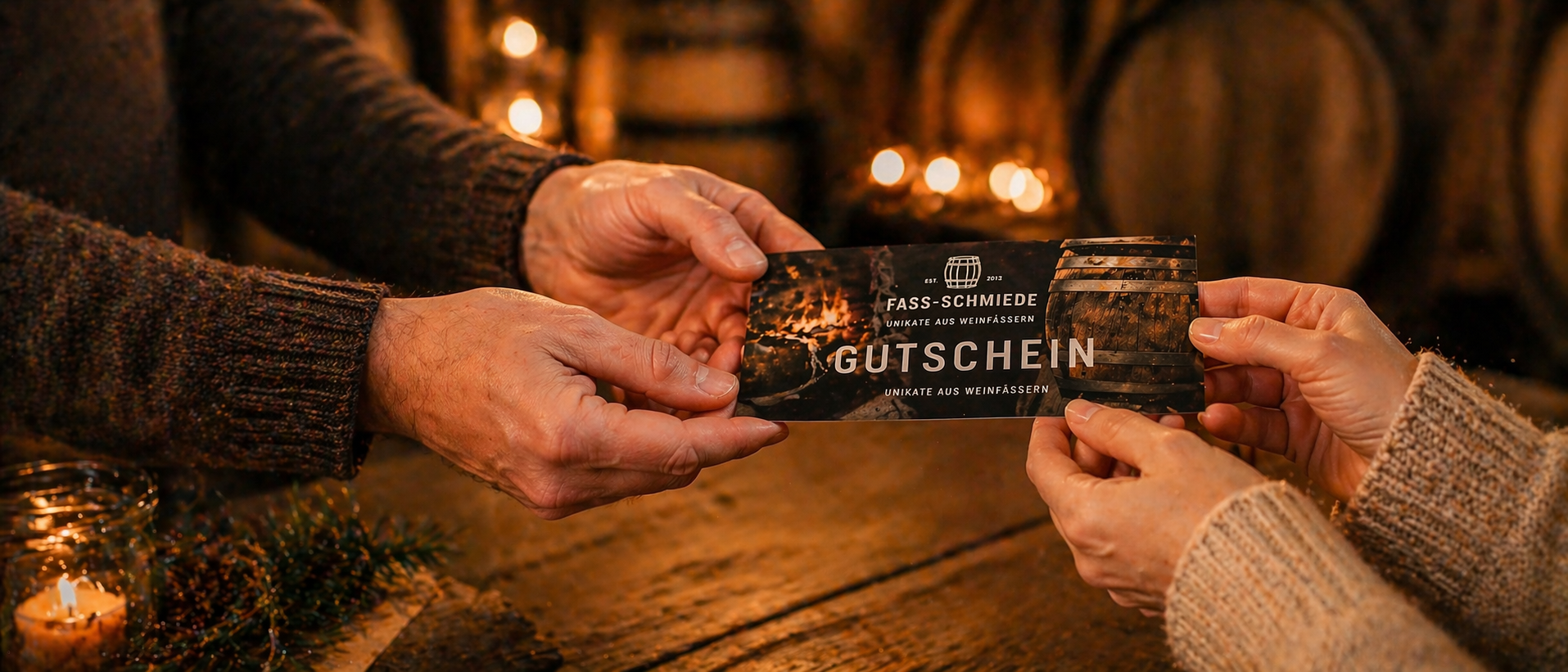 Zwei Hände überreichen einen hochwertigen Gutschein der Fass Schmiede in warmem, stimmungsvollem Licht; im Hintergrund sorgen Kerzen und Holzdetails für eine gemütliche, emotionale Atmosphäre.
