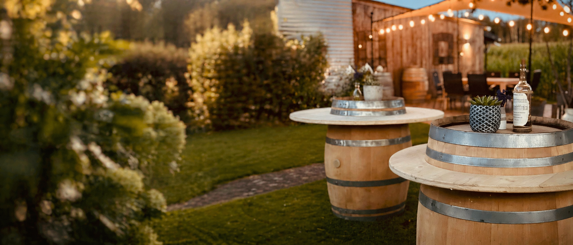 Weinfass Stehtische aus Holz im Garten bei Abendlicht, rechts im Bild arrangiert, mit grünem Garten und rustikalem Schuppen im Hintergrund