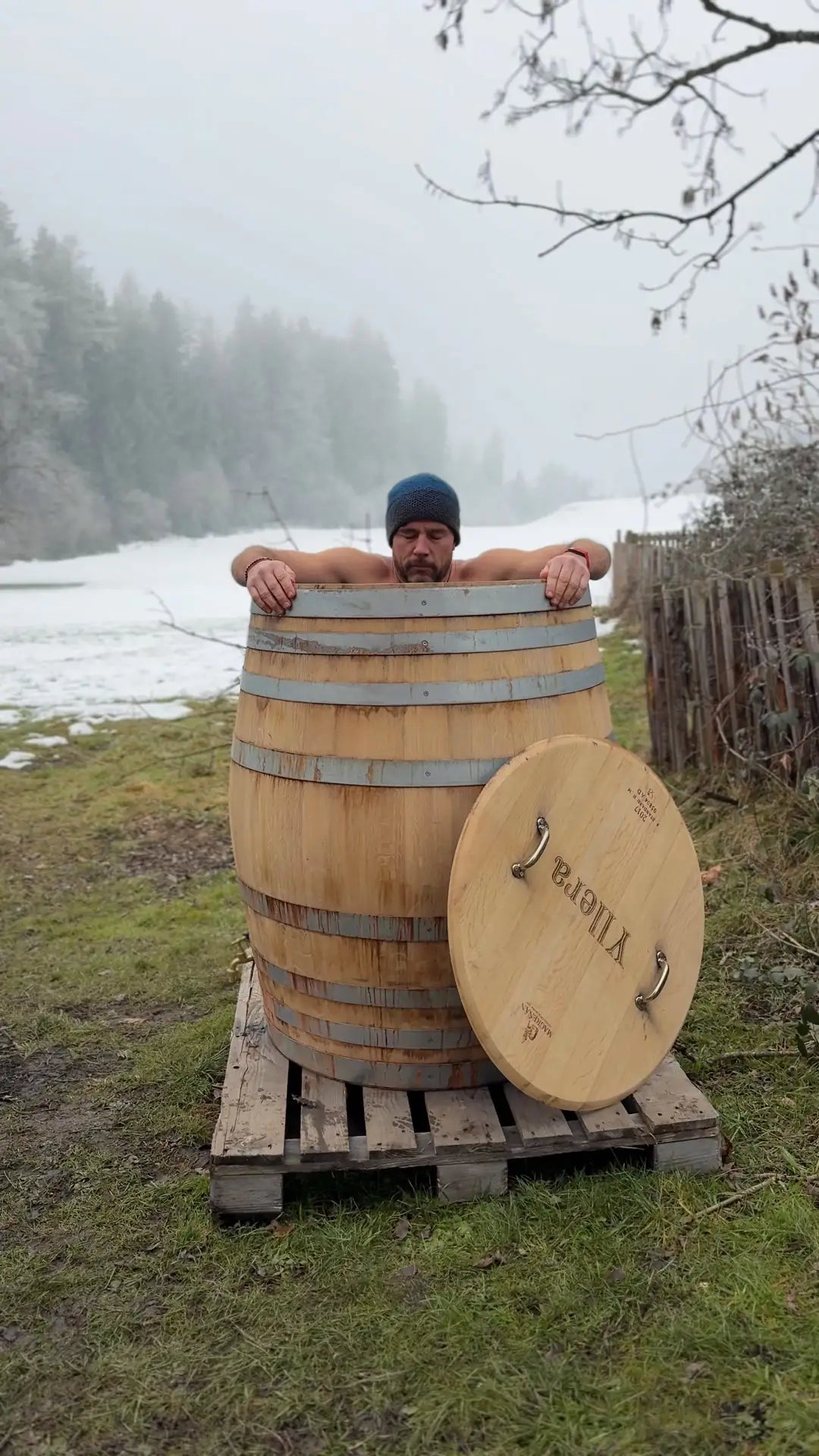 Mann beim Eisbaden in einem Holzfass im Freien vor einer verschneiten Landschaft.